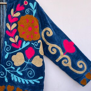 May include: A blue velvet jacket with colorful floral embroidery. The jacket features pink, gold, and turquoise embroidered flowers and leaves. Brown heart shapes are embroidered along the bottom hem. The jacket's interior lining has a striped pattern.