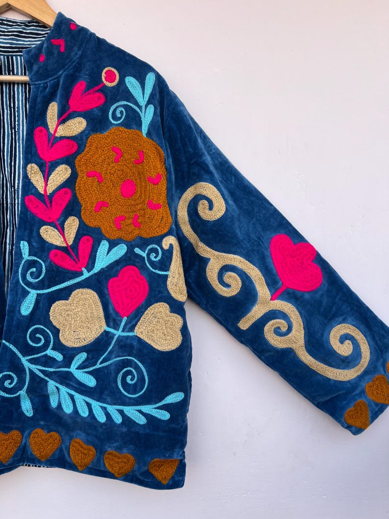 May include: A blue velvet jacket with colorful floral embroidery. The jacket features pink, gold, and turquoise embroidered flowers and leaves. Brown heart shapes are embroidered along the bottom hem. The jacket's interior lining has a striped pattern.