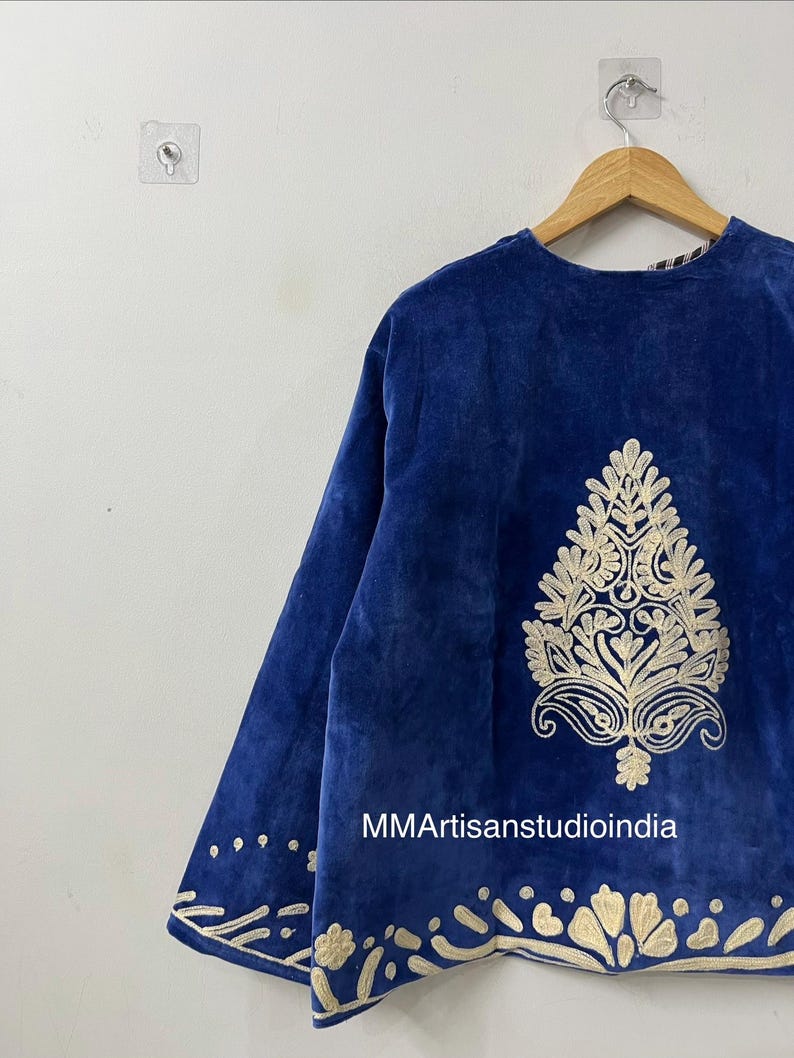May include: A royal blue velvet jacket featuring intricate cream-colored embroidery. The design includes a large central botanical motif on the back and a decorative border along the hem. The text MMArtisanstudioindia is printed on the fabric.