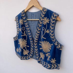 May include: A blue vest with gold floral embroidery. The vest is sleeveless and has a striped lining. It is hanging on a wooden hanger against a white background. The embroidery features detailed floral and leaf patterns.