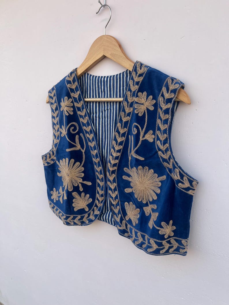 May include: A blue vest with gold floral embroidery. The vest is sleeveless and has a striped lining. It is hanging on a wooden hanger against a white background. The embroidery features detailed floral and leaf patterns.