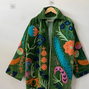 May include: A long-sleeved green velvet coat featuring vibrant, multi-colored floral and botanical embroidery. The coat has a collared neckline, an open front, and a white and green striped lining visible along the inner edges of the garment.