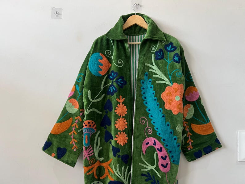 May include: A long-sleeved green velvet coat featuring vibrant, multi-colored floral and botanical embroidery. The coat has a collared neckline, an open front, and a white and green striped lining visible along the inner edges of the garment.