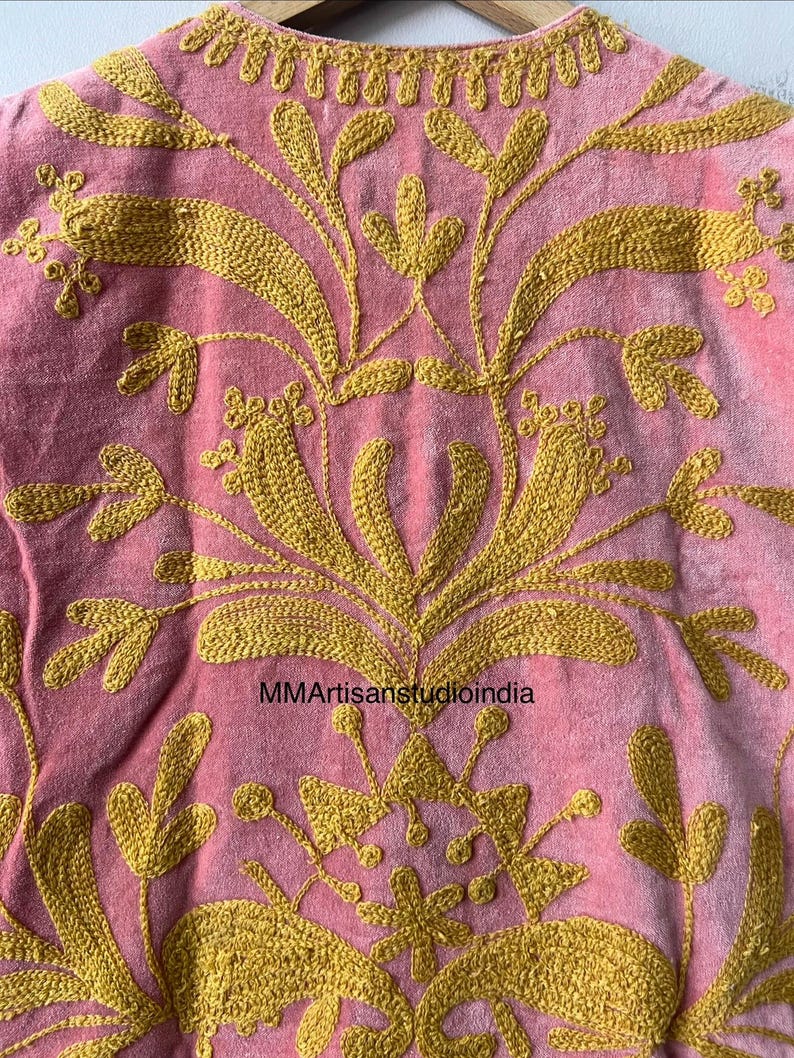 May include: Close-up of a pink textile with intricate gold embroidery. The design features floral and leaf patterns, with the text "MMArtisanstudioindia" visible. The embroidery is detailed and covers most of the fabric.