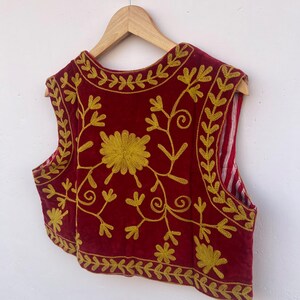 May include: A red sleeveless vest with gold floral embroidery. The vest is displayed on a wooden hanger. The embroidery features a large central flower and detailed vines and leaves. The background is white.