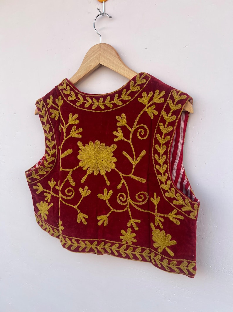 May include: A red sleeveless vest with gold floral embroidery. The vest is displayed on a wooden hanger. The embroidery features a large central flower and detailed vines and leaves. The background is white.
