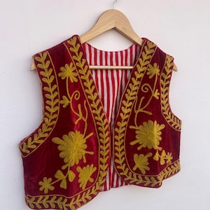 May include: A burgundy velvet vest with gold floral embroidery. The vest has a striped red and white lining and is displayed on a wooden hanger. The embroidery features detailed floral and leaf patterns, creating a decorative garment.