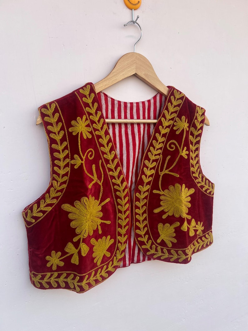 May include: A burgundy velvet vest with gold floral embroidery. The vest has a striped red and white lining and is displayed on a wooden hanger. The embroidery features detailed floral and leaf patterns, creating a decorative garment.