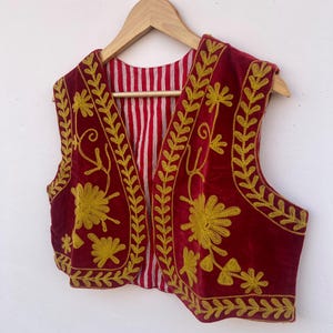May include: A burgundy velvet vest featuring gold floral embroidery and leaf patterns. The vest has a red and white striped lining and is displayed on a wooden hanger. The embroidery is detailed and covers the front and edges.
