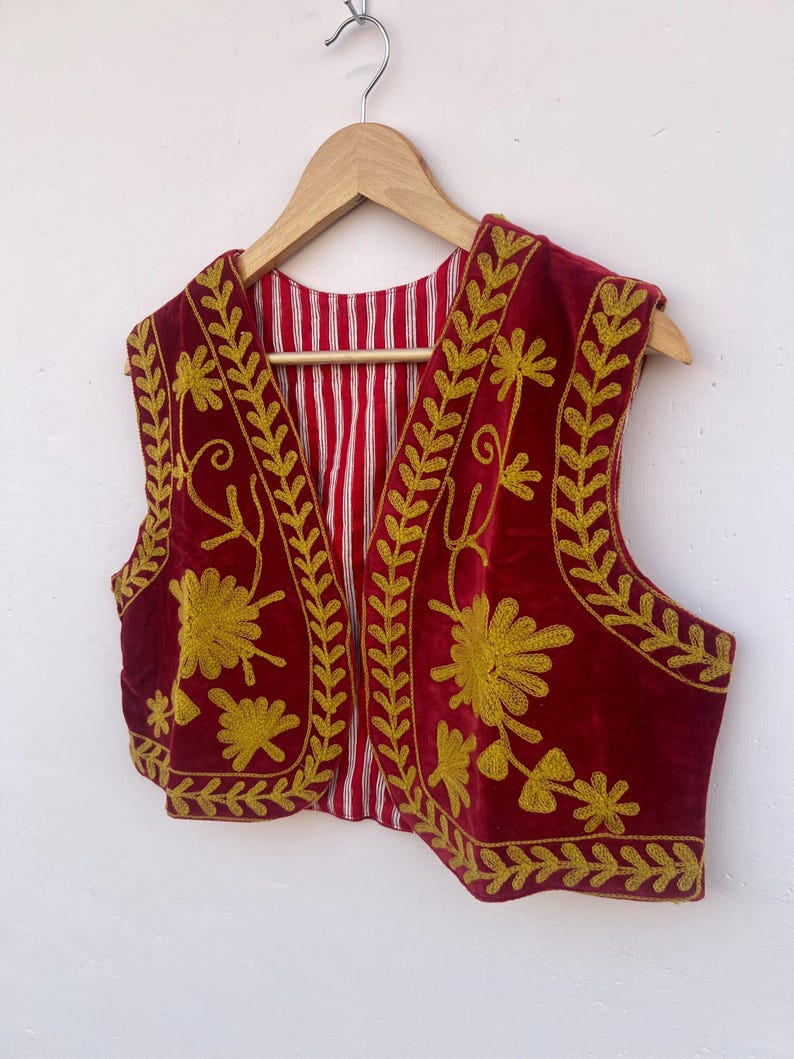 May include: A burgundy velvet vest featuring gold floral embroidery and leaf patterns. The vest has a red and white striped lining and is displayed on a wooden hanger. The embroidery is detailed and covers the front and edges.