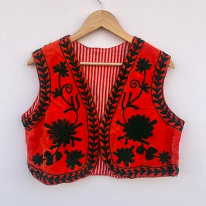 May include: A red vest with black floral embroidery and a striped lining. The vest is hanging on a wooden hanger against a white background. The embroidery features detailed floral patterns.