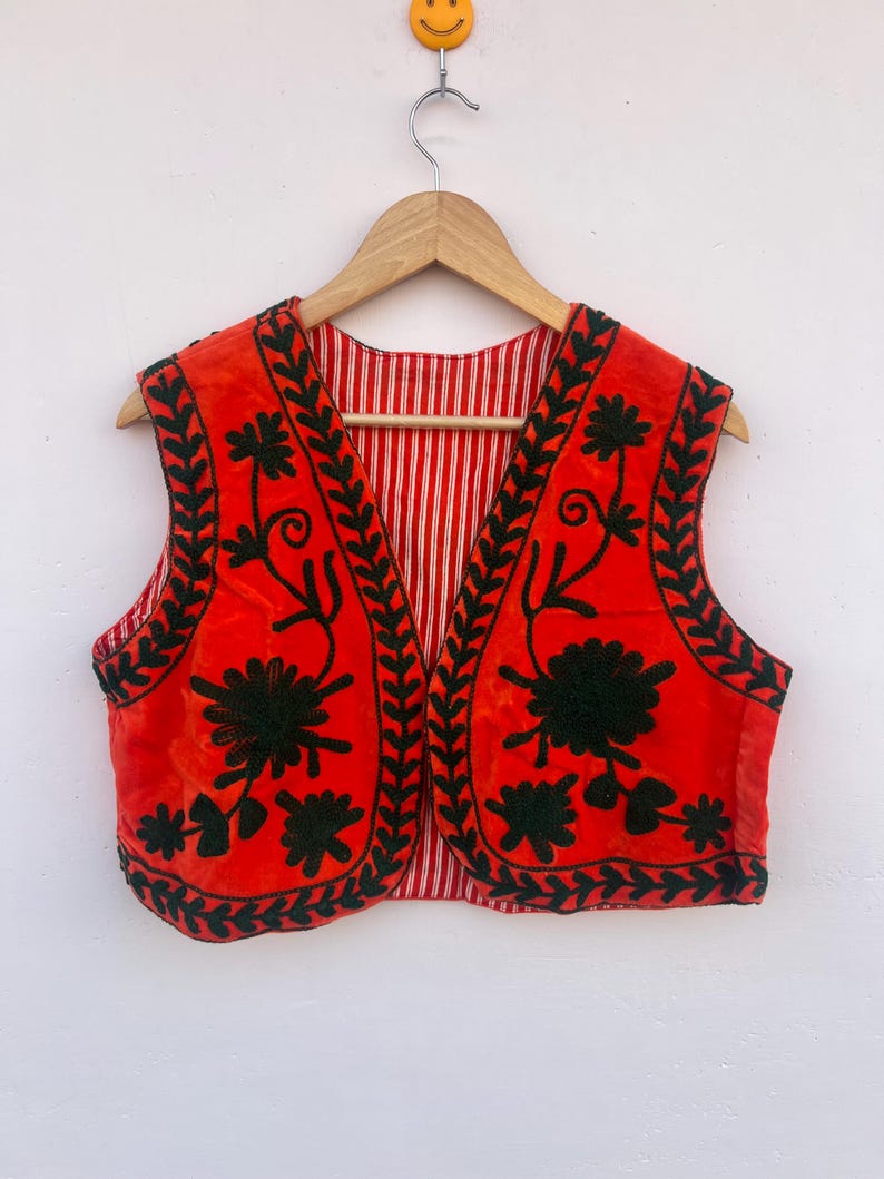 May include: A red vest with black floral embroidery and a striped lining. The vest is hanging on a wooden hanger against a white background. The embroidery features detailed floral patterns.