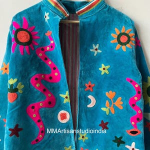 May include: Turquoise velvet jacket with colorful embroidered floral and abstract designs. Features a striped collar and lining. The jacket is adorned with suns, stars, and a pink snake-like design. The text "MMArtisanstudioindia" is visible.
