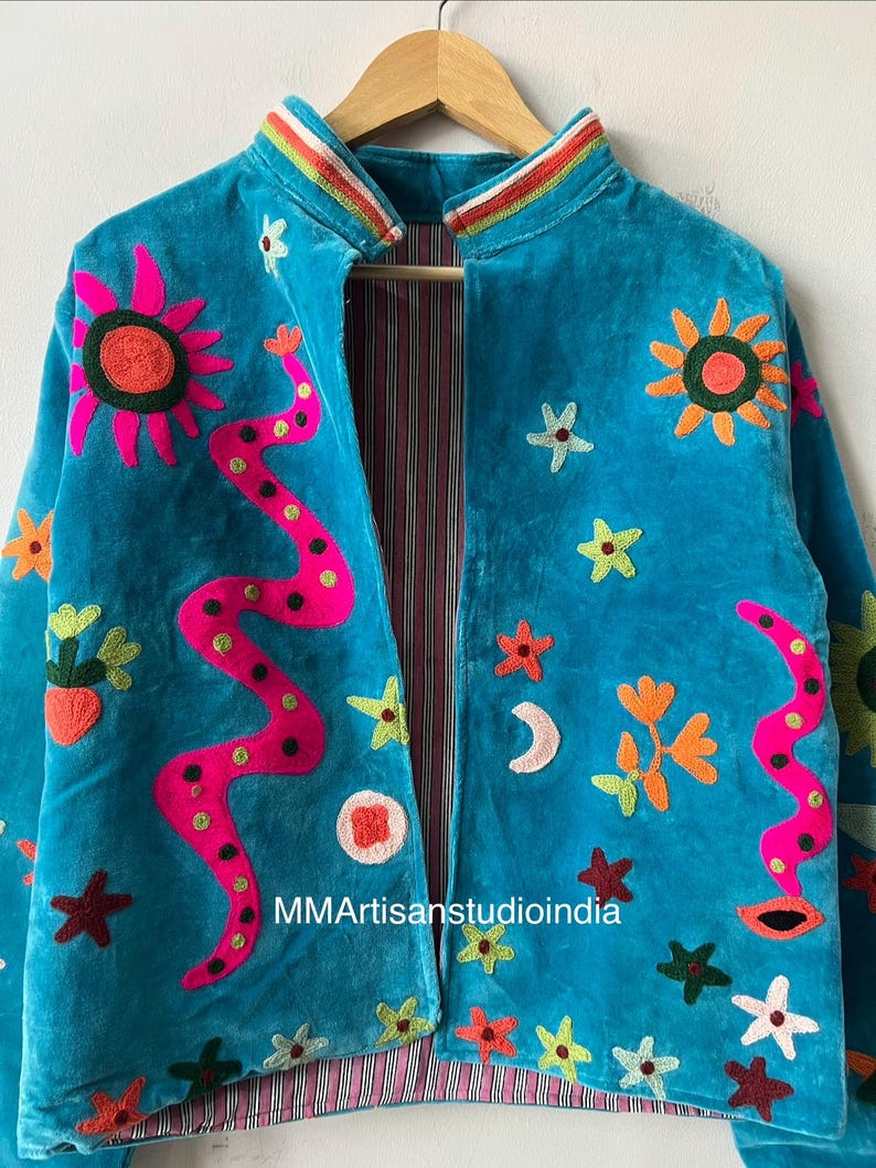 May include: Turquoise velvet jacket with colorful embroidered floral and abstract designs. Features a striped collar and lining. The jacket is adorned with suns, stars, and a pink snake-like design. The text "MMArtisanstudioindia" is visible.