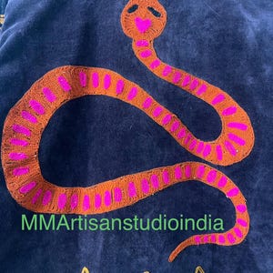 Puede incluir: Tela azul oscuro con un dise&ntilde;o de serpiente naranja bordada con detalles en rosa. La cabeza de la serpiente tiene una boca en forma de coraz&oacute;n. El texto "MMArtisanstudioindia" es visible. Bordado de estrellas doradas en la parte inferior.