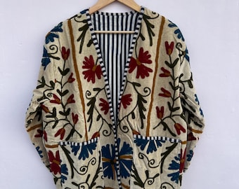 May include: Cream-coloured embroidered jacket with red, blue, and brown floral patterns. The jacket has two pockets and a striped interior. It is hanging on a wooden hanger against a white background.