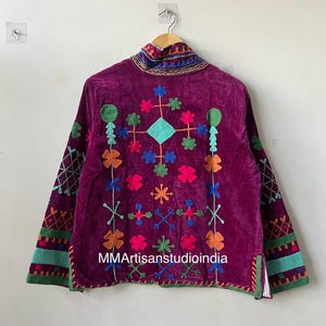 May include: A deep purple velvet jacket with colorful embroidered floral and geometric patterns. The jacket features long sleeves with embroidered cuffs and a high collar. The text "MMArtisanstudioindia" is visible on the front.