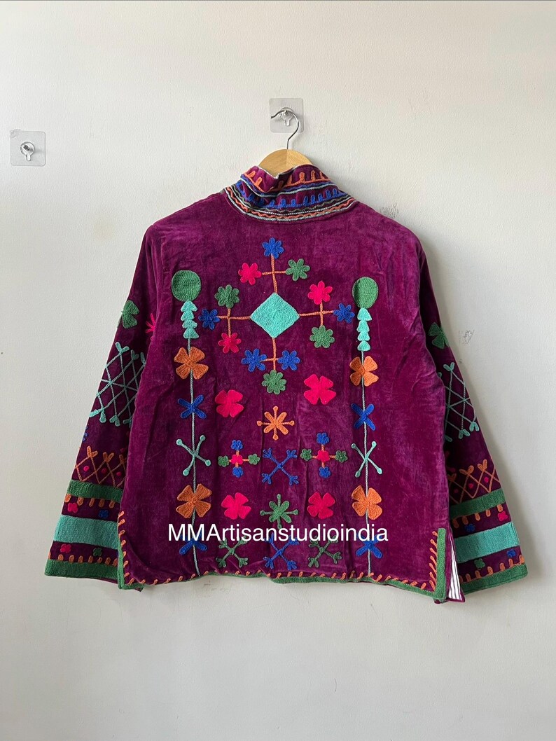 May include: A deep purple velvet jacket with colorful embroidered floral and geometric patterns. The jacket features long sleeves with embroidered cuffs and a high collar. The text "MMArtisanstudioindia" is visible on the front.