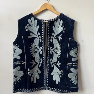 Puede incluir: Un chaleco azul marino con intrincados bordados florales en gris claro. El chaleco sin mangas presenta un dise&ntilde;o detallado en la parte delantera y trasera, con un escote sencillo y una abertura de largo completo. El bordado es una caracter&iacute;stica destacada.