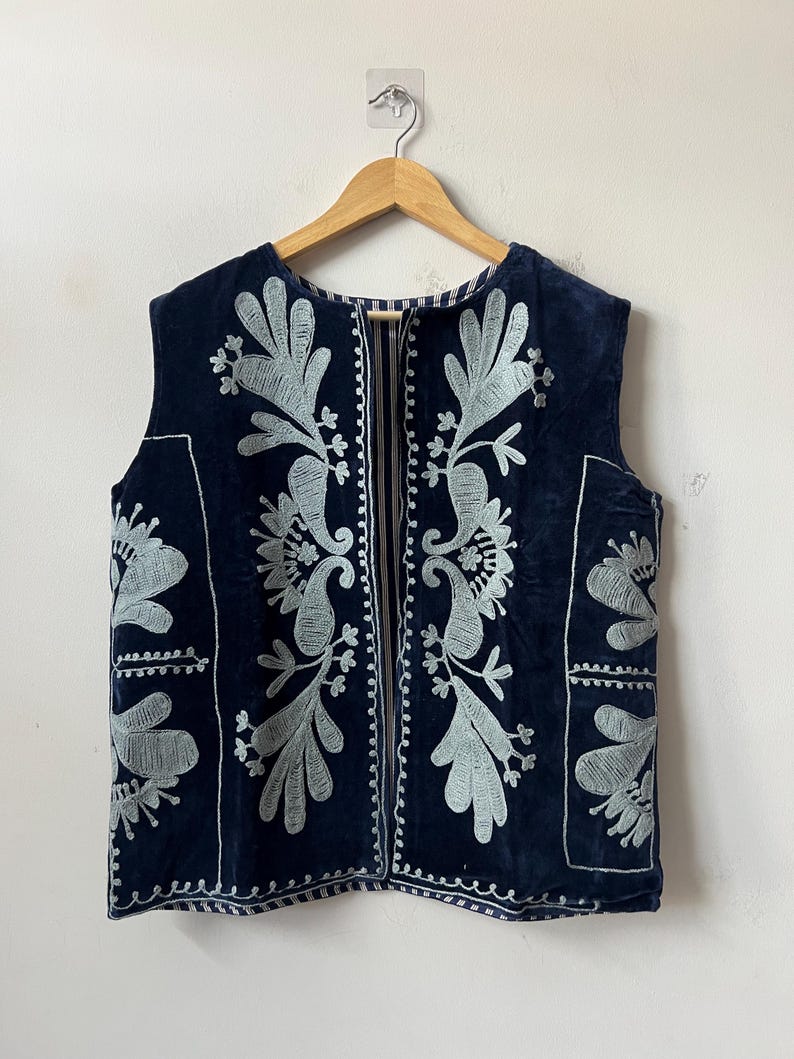 Puede incluir: Un chaleco azul marino con intrincados bordados florales en gris claro. El chaleco sin mangas presenta un dise&ntilde;o detallado en la parte delantera y trasera, con un escote sencillo y una abertura de largo completo. El bordado es una caracter&iacute;stica destacada.
