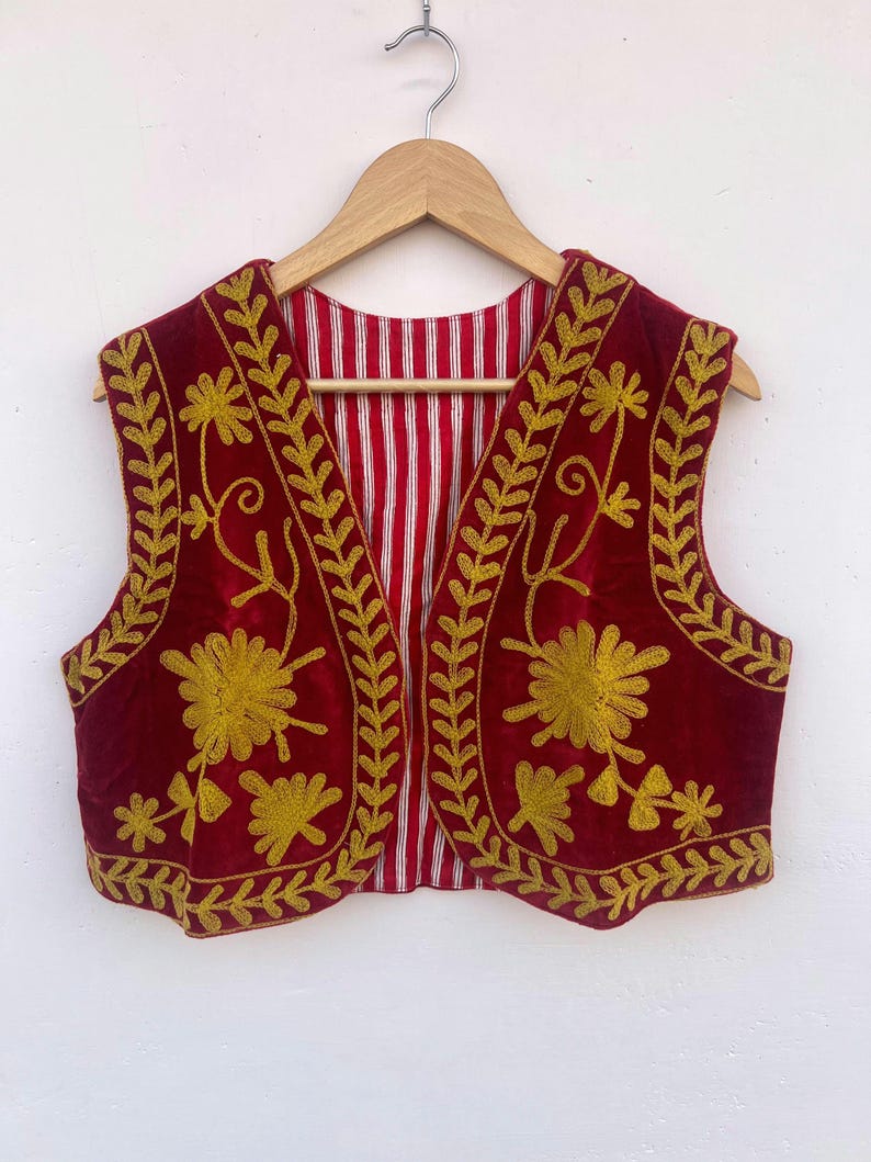 May include: A burgundy vest with gold floral embroidery and a red and white striped lining. The vest is displayed on a wooden hanger. The background is white.