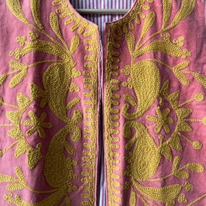 May include: A pink, open-front vest with intricate gold embroidery. The embroidery features floral and paisley designs. The vest's interior lining is a striped pattern of white and pink. The vest is hanging on a wooden hanger.