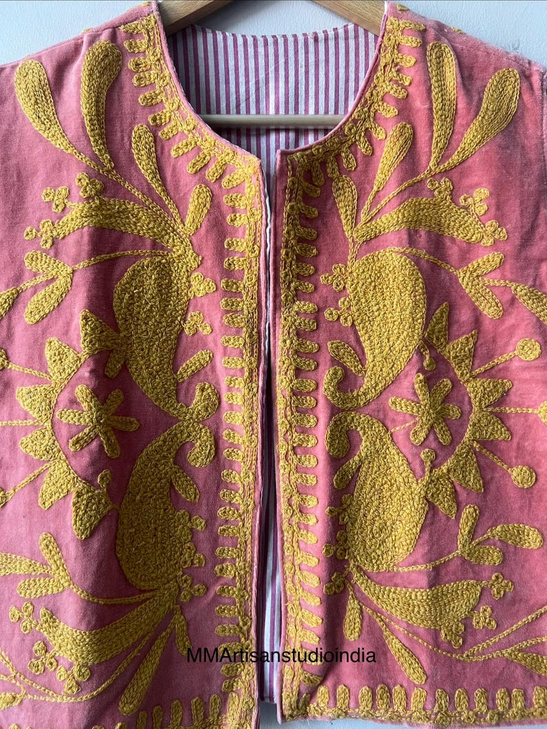 May include: A pink, open-front vest with intricate gold embroidery. The embroidery features floral and paisley designs. The vest's interior lining is a striped pattern of white and pink. The vest is hanging on a wooden hanger.