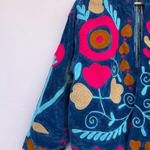 May include: A blue velvet jacket with vibrant floral embroidery. The jacket features a stand-up collar and long sleeves. The embroidery includes pink, gold, and turquoise flowers and leaves. The jacket's interior lining has a striped pattern.
