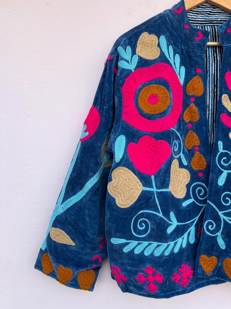 May include: A blue velvet jacket with vibrant floral embroidery. The jacket features a stand-up collar and long sleeves. The embroidery includes pink, gold, and turquoise flowers and leaves. The jacket's interior lining has a striped pattern.
