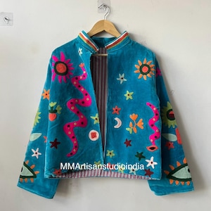 May include: Teal velvet jacket with colorful embroidered celestial and floral designs. Features a pink snake, suns, moons, and stars. The jacket has a striped lining and the text "MMArtisanstudioindia" is visible.