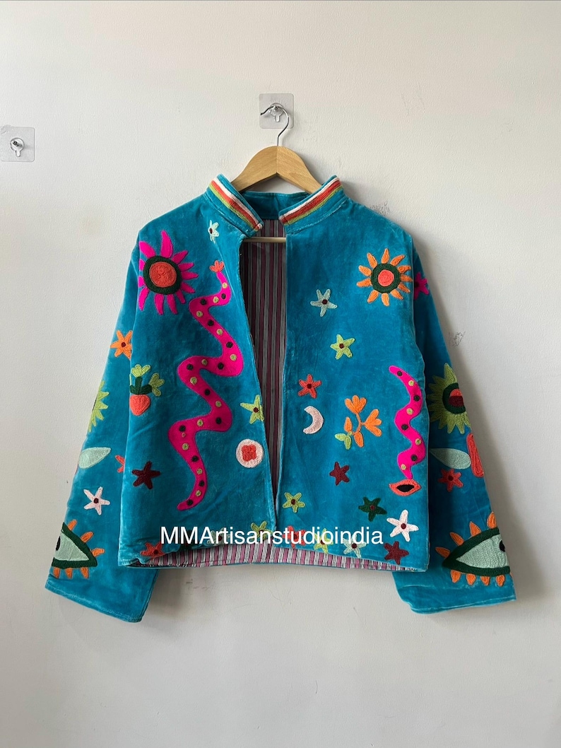 May include: Teal velvet jacket with colorful embroidered celestial and floral designs. Features a pink snake, suns, moons, and stars. The jacket has a striped lining and the text "MMArtisanstudioindia" is visible.