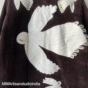 May include: A dark brown jacket with white embroidered bird designs. The jacket features a detailed floral pattern at the hemline. The text "MMArtisanstudioindia" is visible on the lower left side. This handcrafted jacket is a unique piece of apparel.