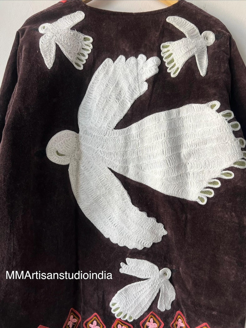May include: A dark brown jacket with white embroidered bird designs. The jacket features a detailed floral pattern at the hemline. The text "MMArtisanstudioindia" is visible on the lower left side. This handcrafted jacket is a unique piece of apparel.