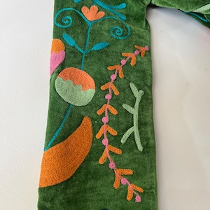 May include: A green velvet sleeve featuring colorful embroidery of stylized flowers, leaves, and vines in shades of orange, pink, light green, turquoise, and dark blue. The fabric has a soft, textured pile, and the stitching is raised and detailed.