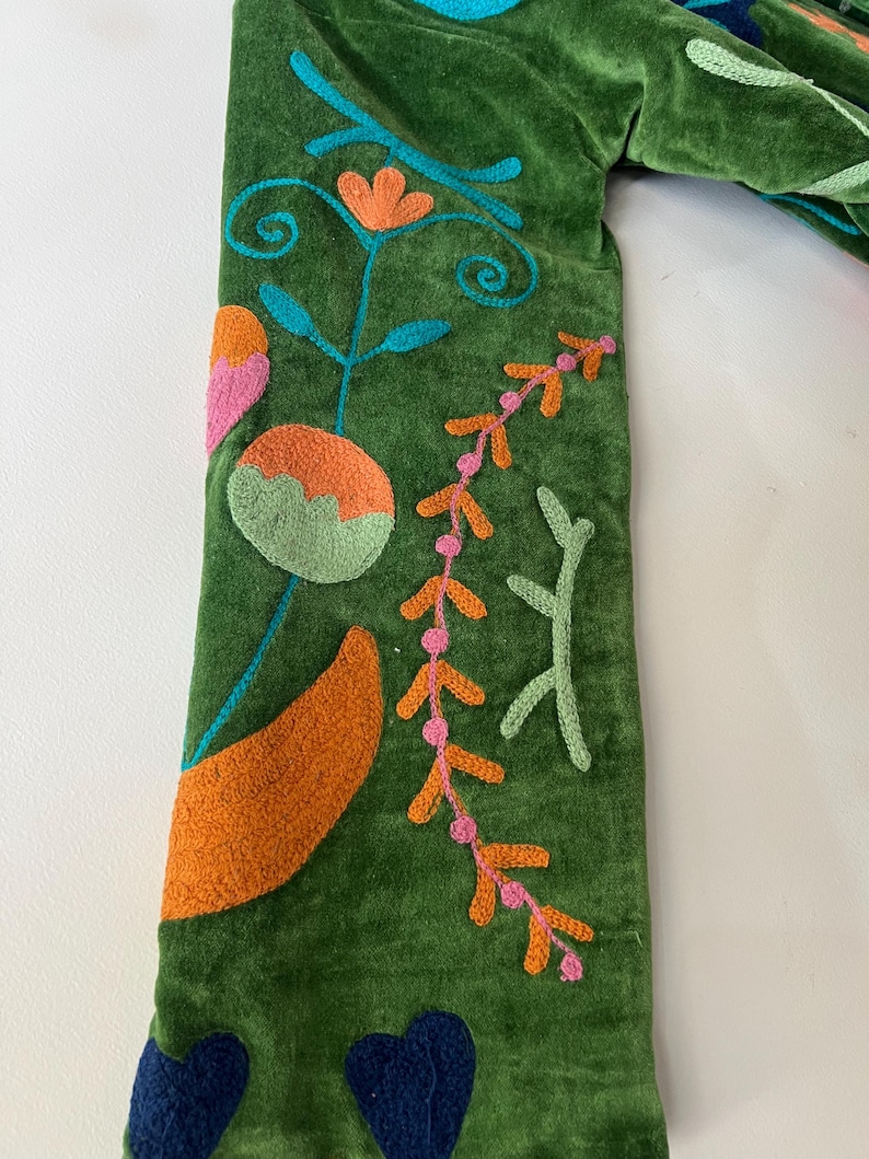 May include: A green velvet sleeve featuring colorful embroidery of stylized flowers, leaves, and vines in shades of orange, pink, light green, turquoise, and dark blue. The fabric has a soft, textured pile, and the stitching is raised and detailed.