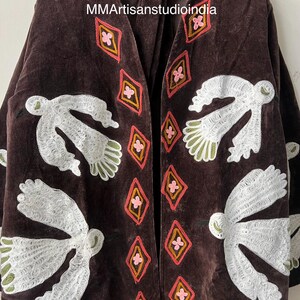May include: A dark brown velvet jacket with white embroidered bird and geometric designs. The jacket features a V-neck and long sleeves. The geometric designs are in pink, orange, and yellow.