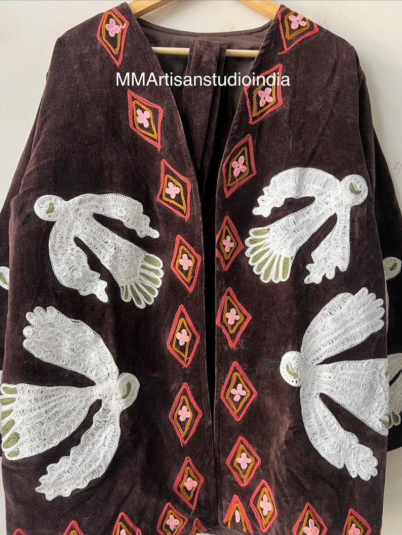 May include: A dark brown velvet jacket with white embroidered bird and geometric designs. The jacket features a V-neck and long sleeves. The geometric designs are in pink, orange, and yellow.