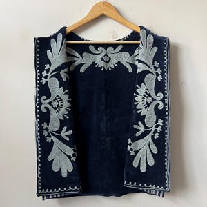 Puede incluir: Un chaleco azul marino con intrincados bordados plateados. El chaleco presenta un detallado dise&ntilde;o floral y de cachemira en los bordes y la espalda. Cuelga de una percha de madera sobre un fondo blanco.