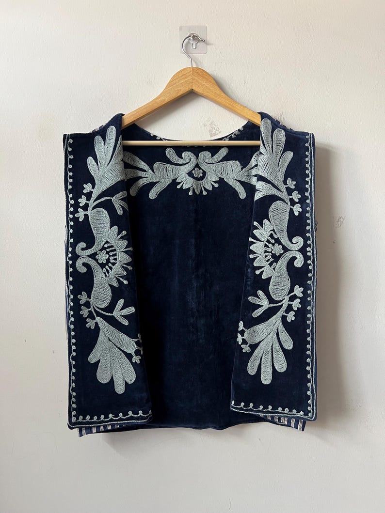Puede incluir: Un chaleco azul marino con intrincados bordados plateados. El chaleco presenta un detallado dise&ntilde;o floral y de cachemira en los bordes y la espalda. Cuelga de una percha de madera sobre un fondo blanco.