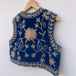 May include: A sleeveless, blue velvet vest with intricate gold floral embroidery. The embroidery includes a large flower in the center and floral patterns along the edges. The vest is hanging on a wooden hanger.