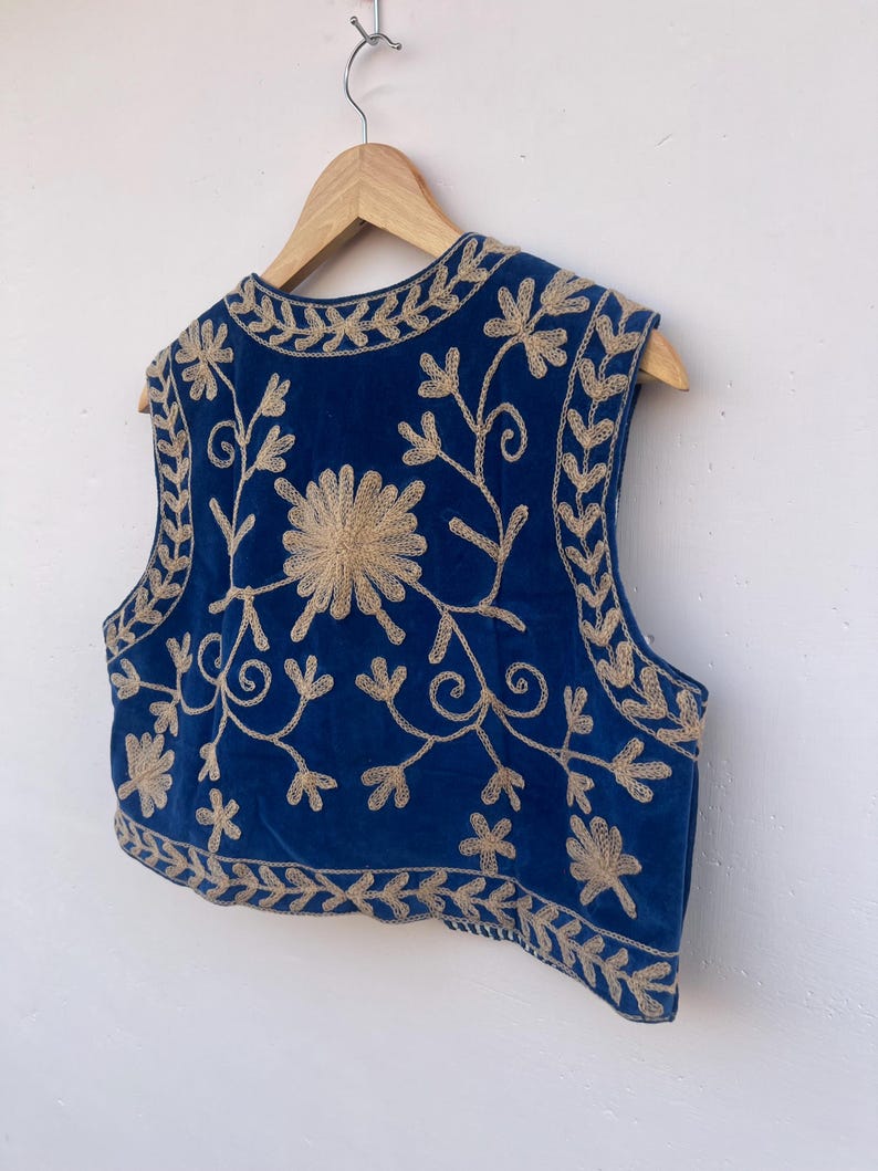 May include: A sleeveless, blue velvet vest with intricate gold floral embroidery. The embroidery includes a large flower in the center and floral patterns along the edges. The vest is hanging on a wooden hanger.