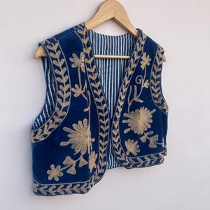 May include: A navy blue vest with gold floral embroidery and a striped lining. The vest is hanging on a wooden hanger. The embroidery is detailed and covers the front and edges of the vest.