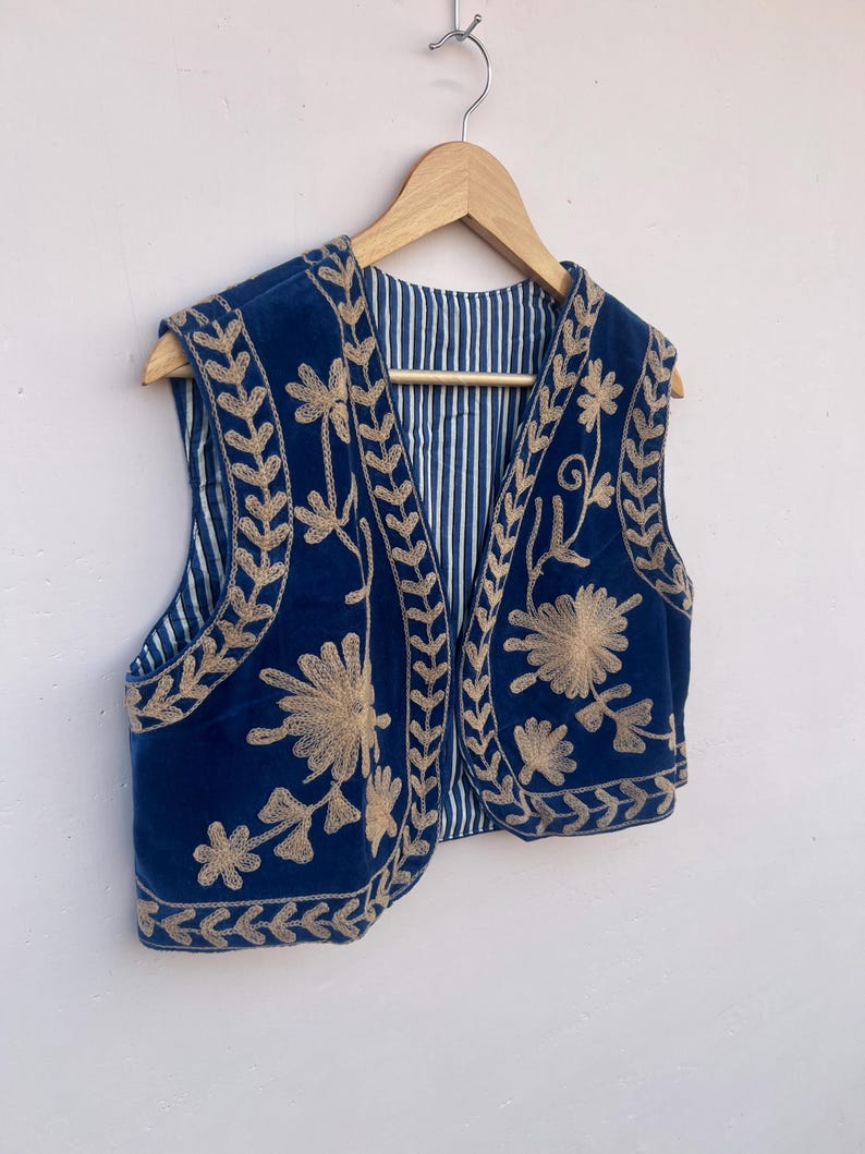 May include: A navy blue vest with gold floral embroidery and a striped lining. The vest is hanging on a wooden hanger. The embroidery is detailed and covers the front and edges of the vest.