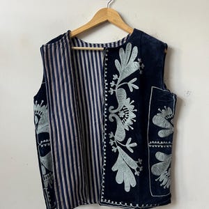Puede incluir: Un chaleco azul marino con interior a rayas y bordados florales. El chaleco sin mangas cuelga de una percha de madera. El bordado es de color azul claro. El chaleco est&aacute; contra una pared blanca.