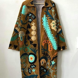 May include: A brown long-sleeve coat featuring colorful embroidered floral and mushroom motifs in shades of turquoise, orange, and black. The coat has a collared neckline, a buttoned cuff detail, and a black and white vertical striped interior lining.