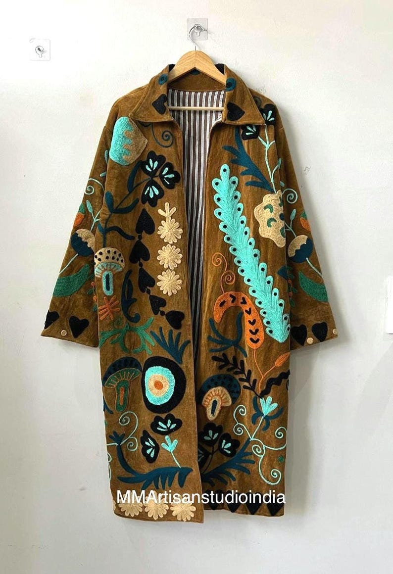 May include: A brown long-sleeve coat featuring colorful embroidered floral and mushroom motifs in shades of turquoise, orange, and black. The coat has a collared neckline, a buttoned cuff detail, and a black and white vertical striped interior lining.