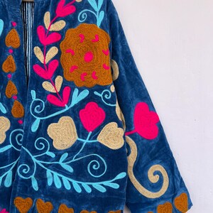 May include: A blue velvet jacket with colorful floral embroidery. The jacket features a stand-up collar and long sleeves. The embroidery includes pink, gold, and turquoise flowers and leaves, with brown heart-shaped accents along the hem.