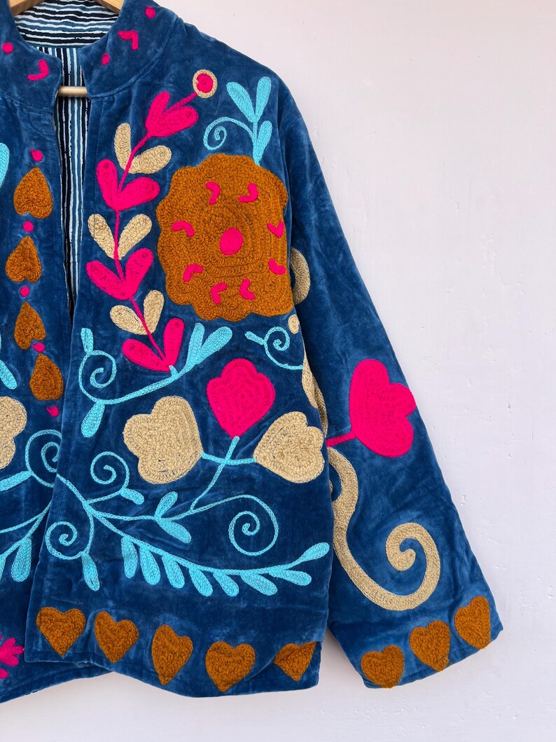 May include: A blue velvet jacket with colorful floral embroidery. The jacket features a stand-up collar and long sleeves. The embroidery includes pink, gold, and turquoise flowers and leaves, with brown heart-shaped accents along the hem.