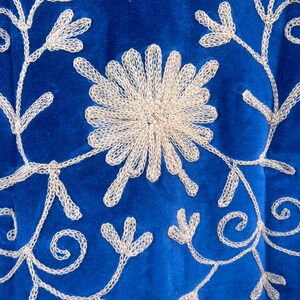 May include: Close-up of a royal blue textile with intricate, cream-colored embroidered floral patterns. The design features a central flower and delicate vines with leaf-like details, creating a textured, decorative surface.