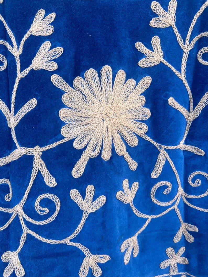 May include: Close-up of a royal blue textile with intricate, cream-colored embroidered floral patterns. The design features a central flower and delicate vines with leaf-like details, creating a textured, decorative surface.